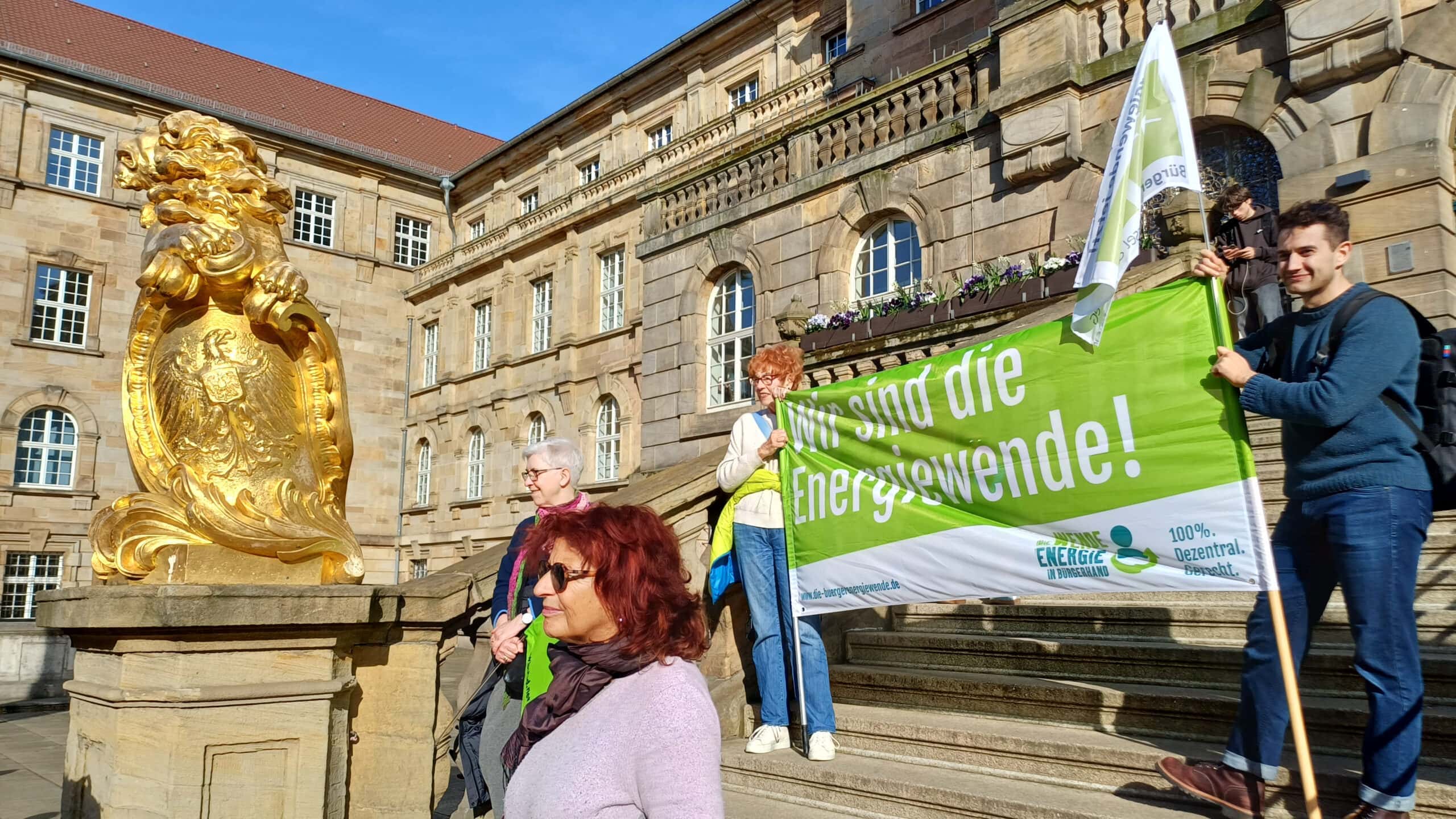 Menschen bei einer Demonstration für erneuerbare Energien vor einem historischen Gebäude mit goldner.